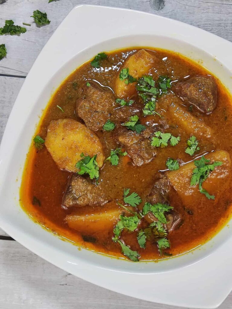 Authentic Aloo Gosht Recipe: Tender Lamb and Potato Curry - Plattershare - Recipes, food stories and food lovers Authentic Indian meat curry with potatoes garnished with fresh cilantro on a white plate.