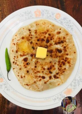 Soft buttery roti with charred spots served on a floral plate, garnished with a green chili and butter cube.