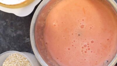 Fresh pink tomato juice in a stainless steel jar with ingredients on a black countertop.