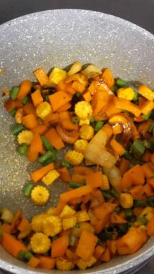 Colorful sautéed mixed vegetables with carrots, baby corn, and onions in a gray non-stick pan.