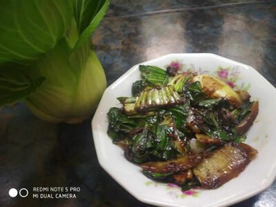 Stir-fried leafy greens and eggplant on a floral dish with fresh vegetables on a dark countertop.