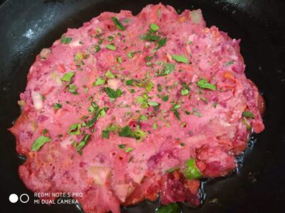 Chopped vegetables cooking into a pink-colored mixture garnished with fresh coriander on a black pan.
