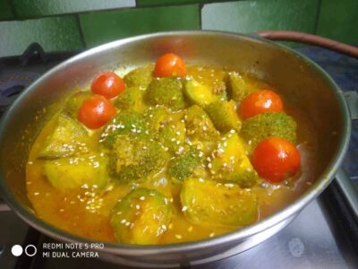 Bright yellow curry with cherry tomatoes, green vegetables, and sesame seeds in a stainless steel pan.